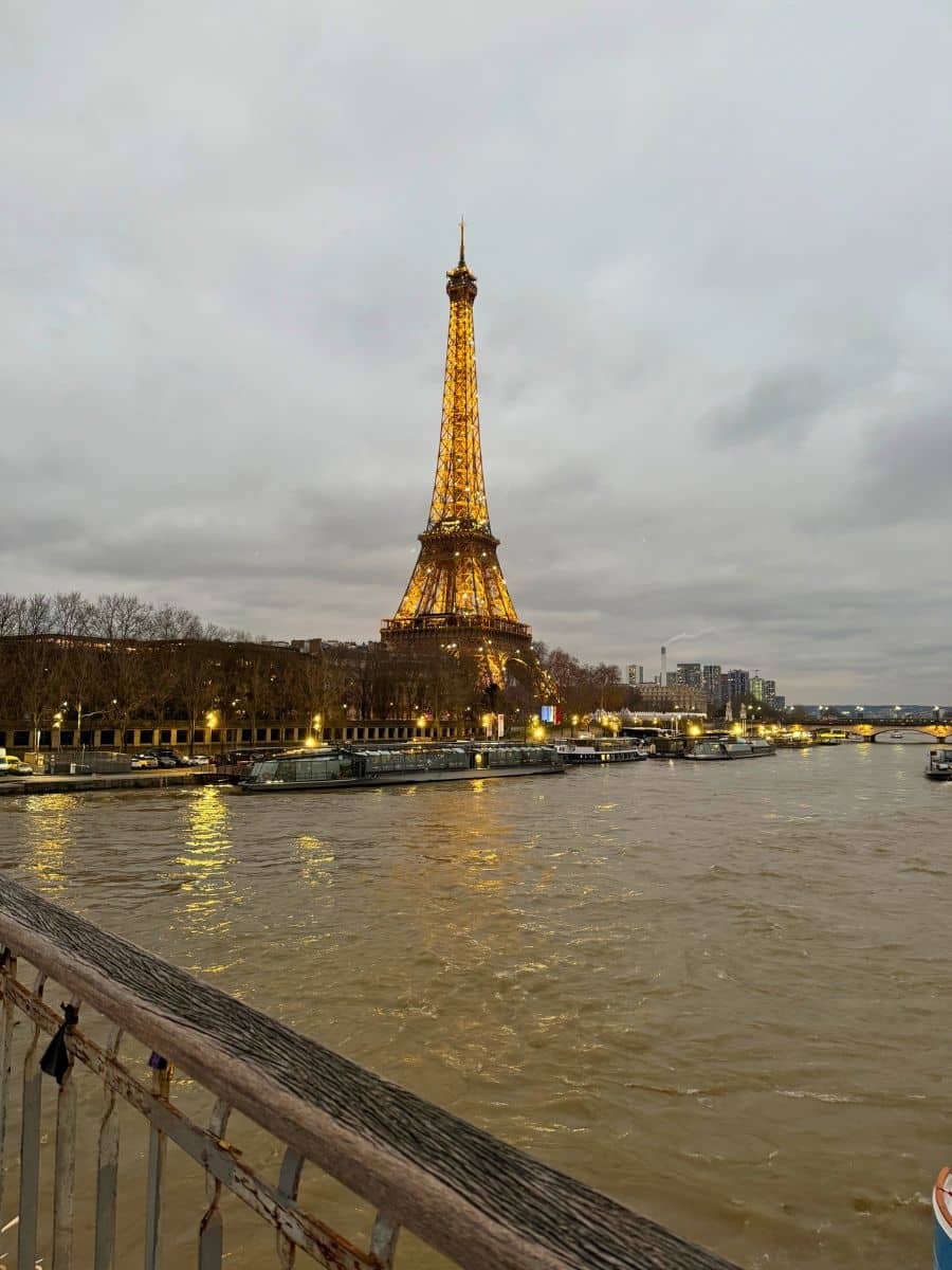 The Eiffel Tower is illuminated against a cloudy evening sky, reflecting on the Seine River. The scene captures a cozy, festive Parisian atmosphere during the Christmas season, with nearby buildings and boats adding to the charm.