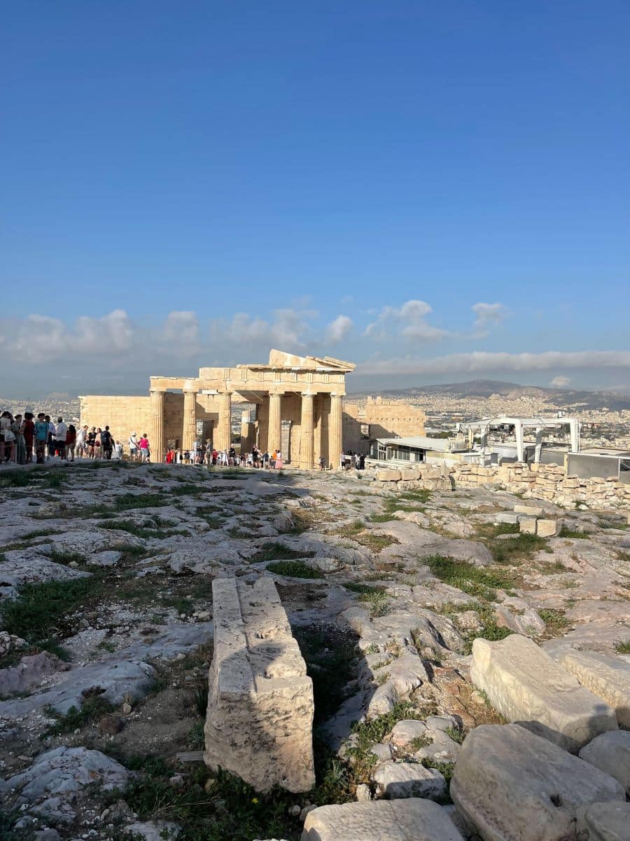 View of the Propylaea, the grand entrance to the Acropolis in Athens, Greece, with tourists exploring the ancient site under a clear blue sky.
