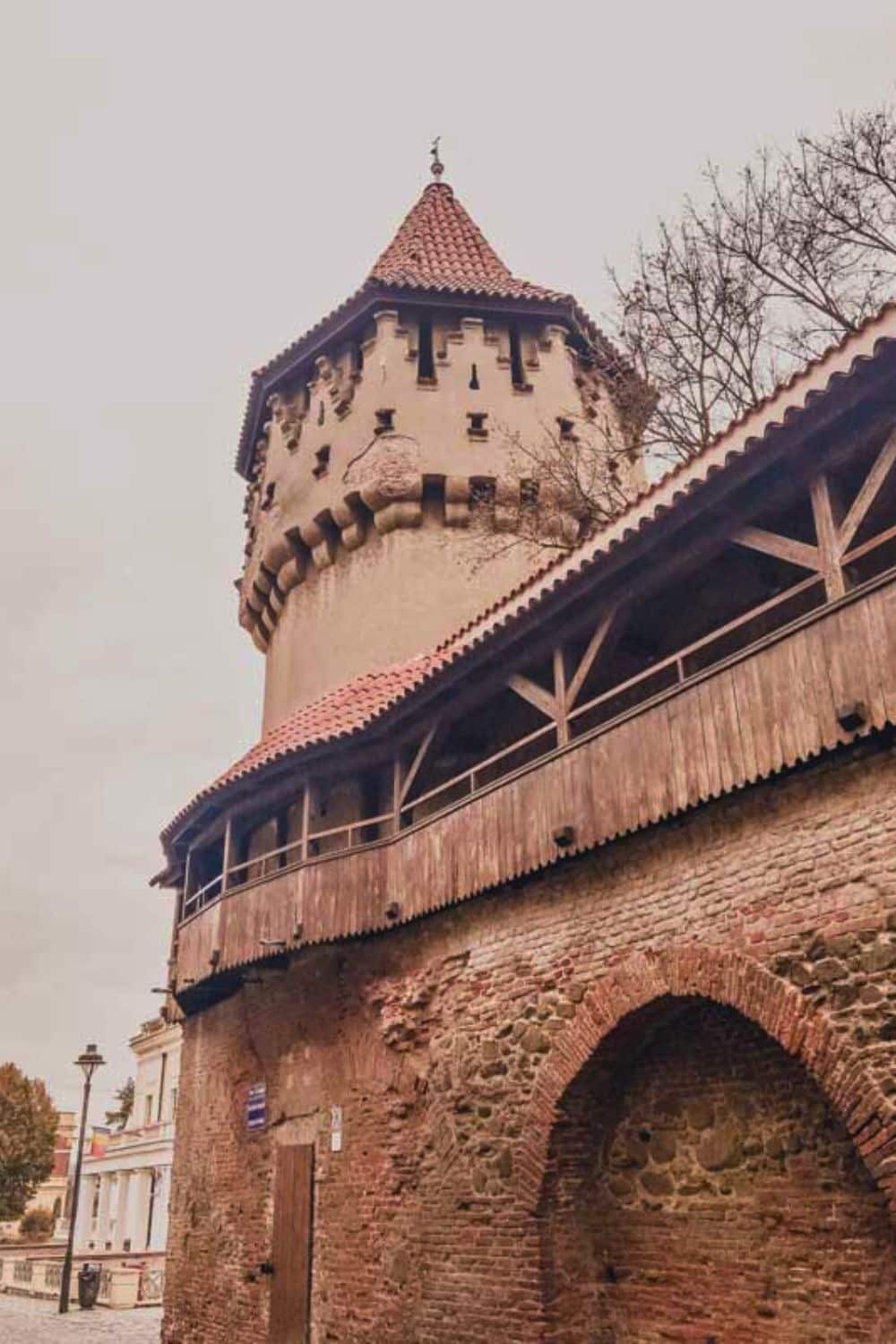 Medieval tower with a conical roof, attached to a section of an old wall with a wooden upper walkway, part of historical fortifications in a European city.