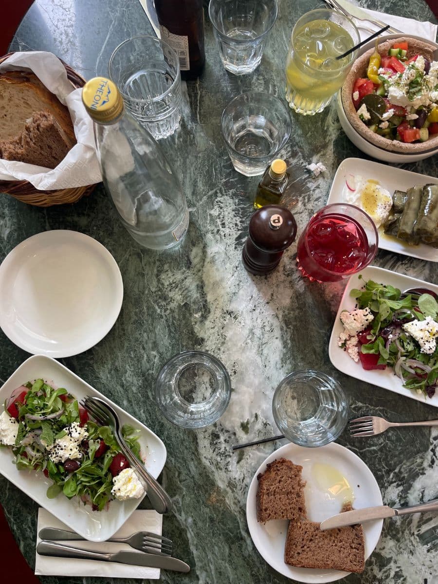 A table set with traditional Greek dishes, including fresh salads with feta cheese, dolmades, bread with olive oil, and various drinks, on a green marble tabletop. The setting suggests a cozy, authentic dining experience.