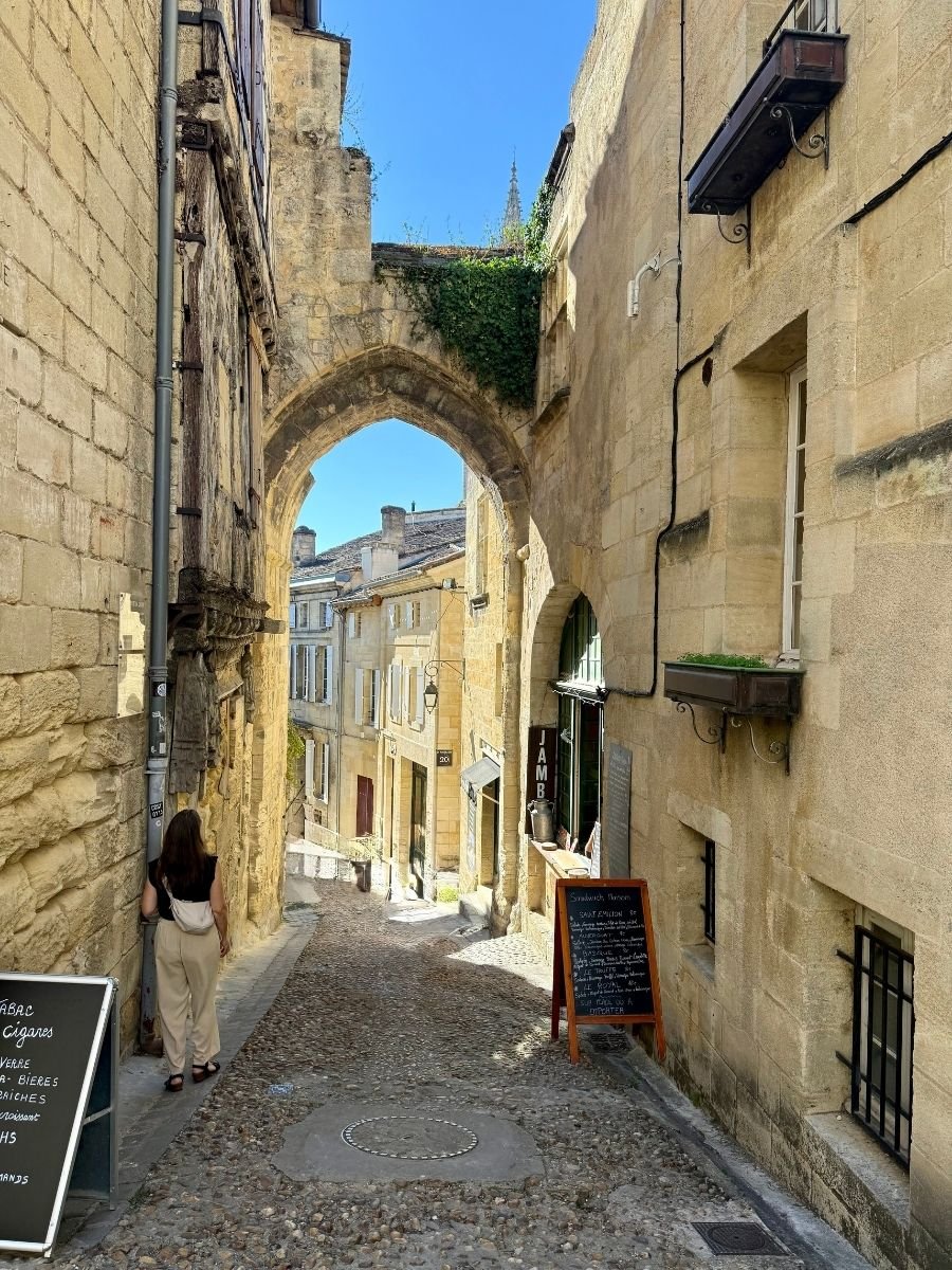 Narrow cobbled alley in Saint-Émilion, Bordeaux, France, framed by a medieval stone archway and lined with shops and chalkboard menus.