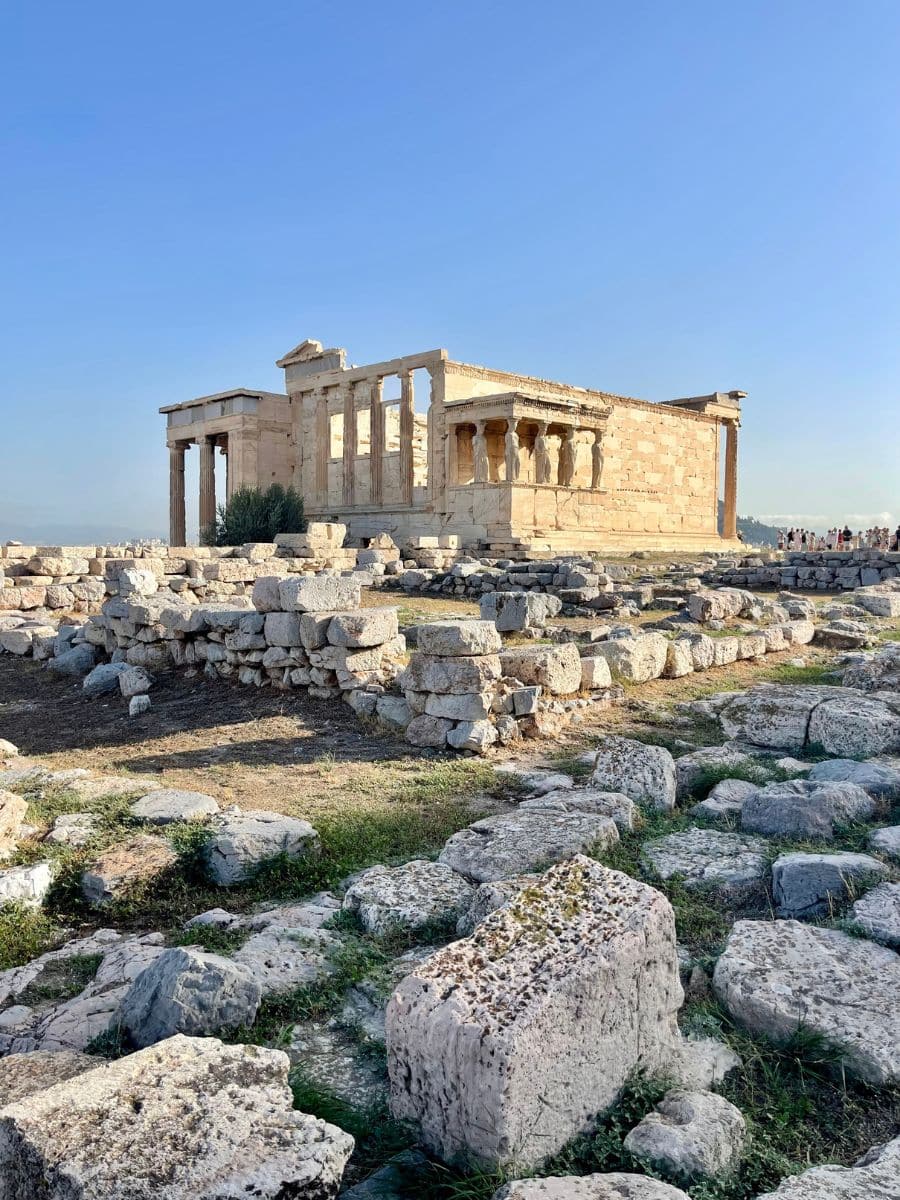The Erechtheion, an ancient Greek temple on the Acropolis of Athens, featuring classical architecture with iconic columns and surrounded by scattered stone ruins under a clear blue sky.