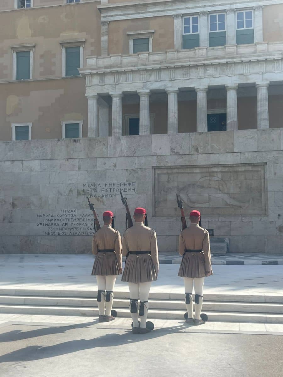 Three Evzones guards in traditional uniform stand in formation in front of the Tomb of the Unknown Soldier in Athens, Greece, outside the Hellenic Parliament.