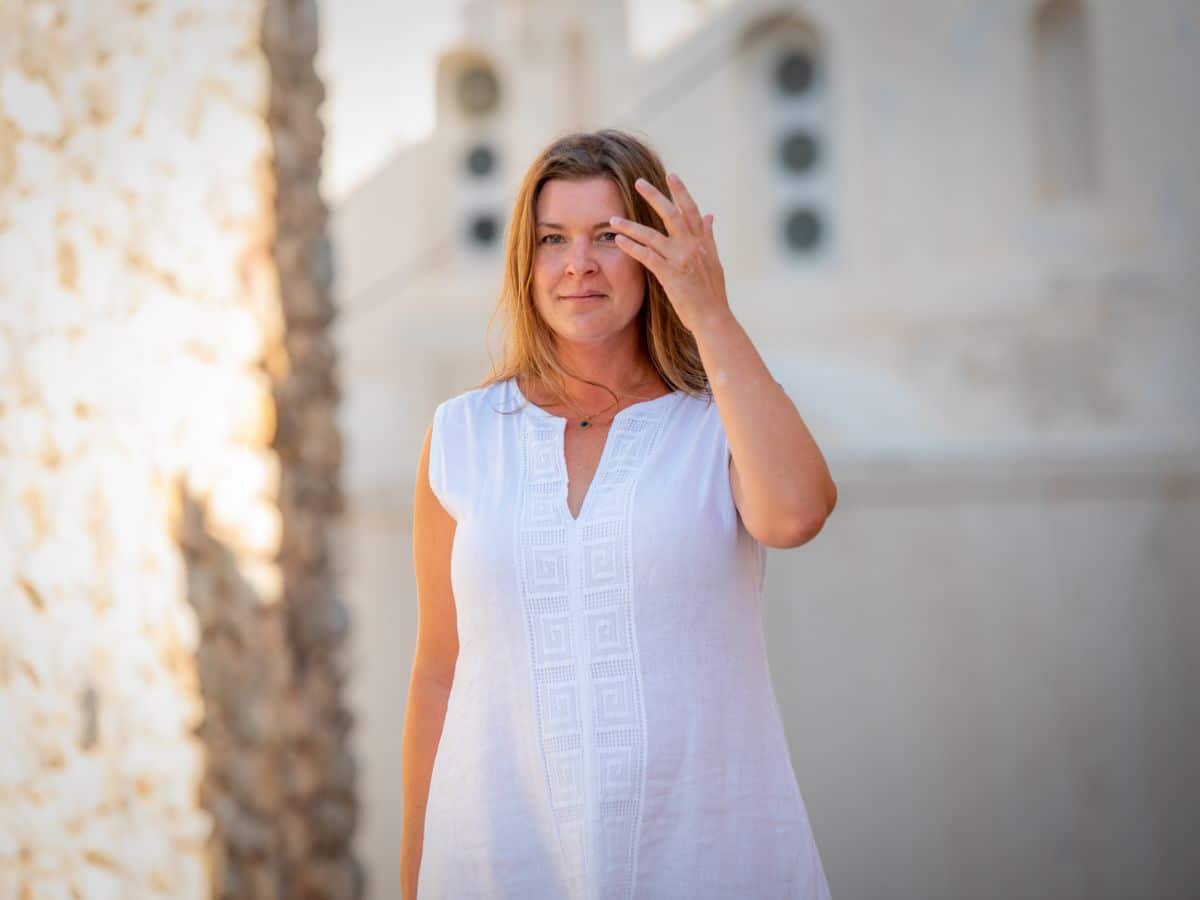 A woman in a white sleeveless dress gently touching her hair, standing in front of a stone building. She is gazing forward with a serene expression.