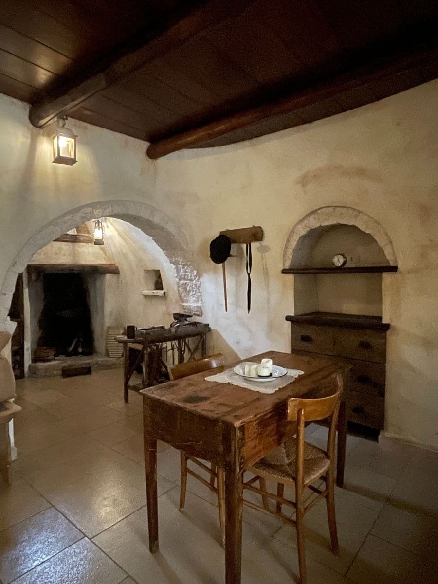 A cozy, rustic interior of a traditional Trullo house in Alberobello, Italy. The room features whitewashed stone walls and a wooden ceiling with exposed beams. A simple wooden dining table with two chairs sits in the center, adorned with a lace placemat and a plate with candles. In the background, an arched alcove houses a fireplace with blackened walls from years of use, along with antique tools. Another smaller arched alcove contains wooden shelves with a vintage clock. A wooden dresser with drawers stands against the wall, contributing to the old-world charm of the space. Soft lighting from a lantern adds warmth to the atmosphere.