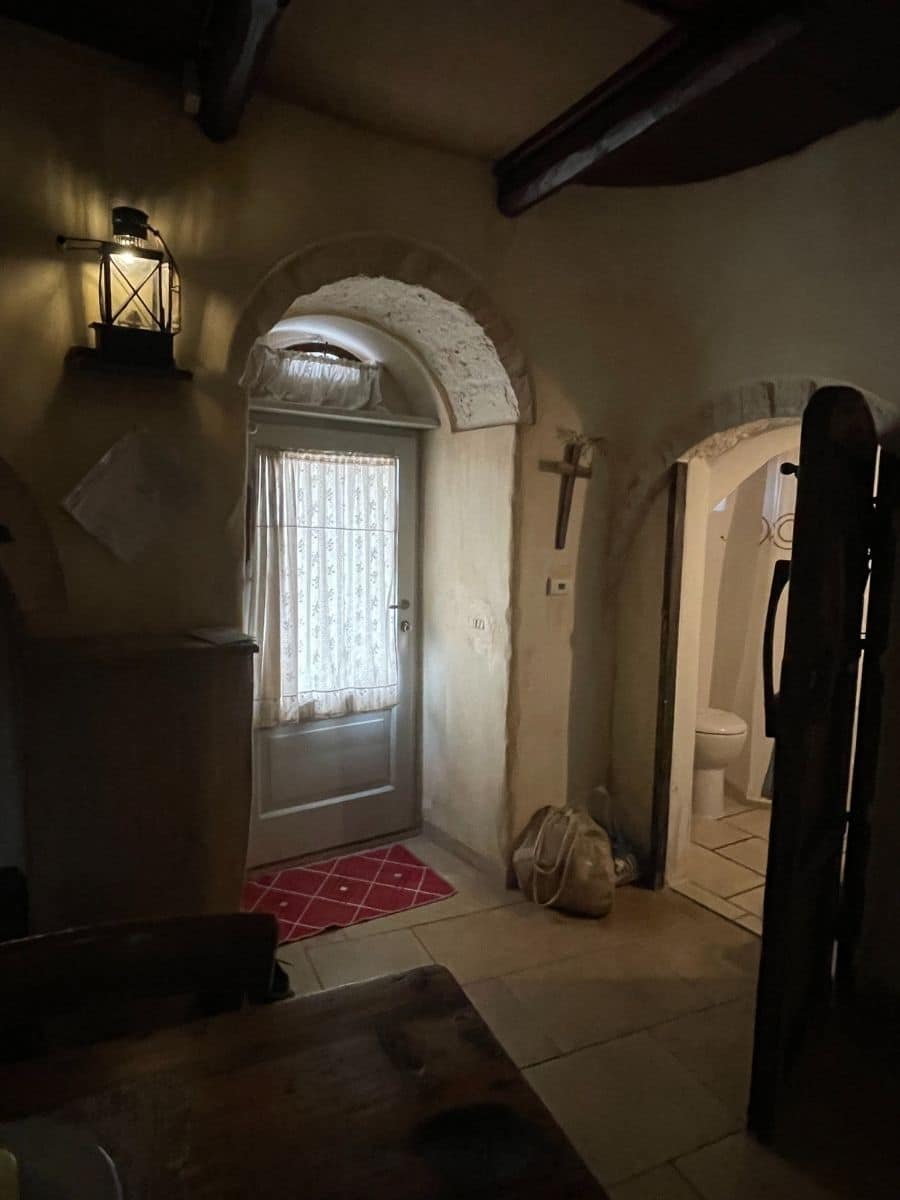 A cozy interior of a traditional Trullo house in Alberobello, Italy. The space features rustic stone walls, arched doorways, and wooden ceiling beams. A soft glow from a lantern-style wall light enhances the warm ambiance. A small doorway leads to a bathroom with a visible toilet. The main door has a white lace curtain, allowing natural light to filter in. A red patterned rug adds a touch of color to the neutral-toned tile floor. A beige canvas bag rests on the floor near the entrance, contributing to the lived-in charm of this historic home.