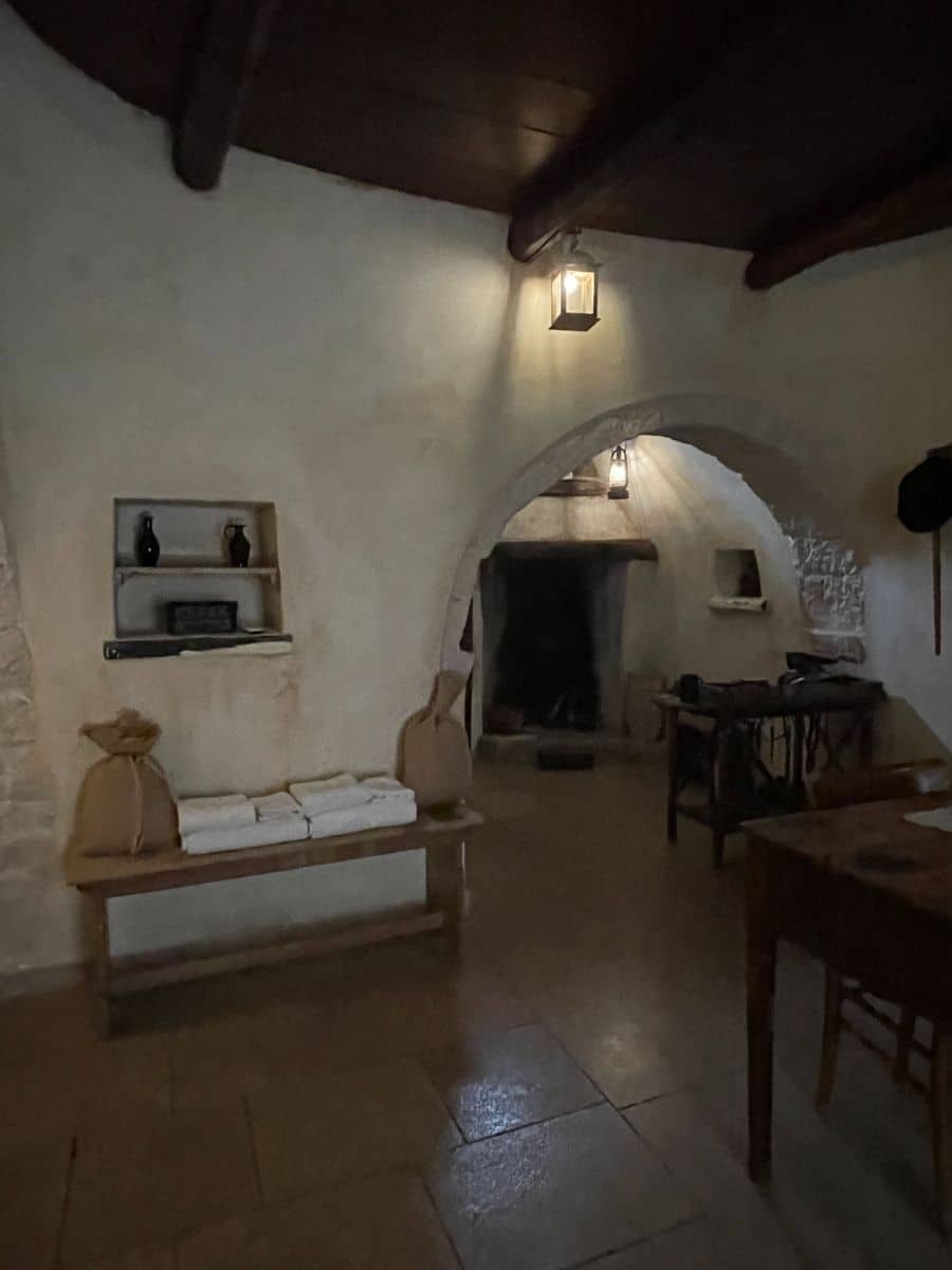 A traditional Trullo house interior in Alberobello, Italy, featuring rustic stone walls and arched doorways. A wooden bench against the wall holds neatly folded white towels and burlap sacks. Above the bench, a small built-in shelf displays black ceramic bottles. A lantern-style wall light casts a warm glow over the space. The arched entrance leads to a dimly lit alcove with a fireplace and a workbench, creating a cozy and historical atmosphere. The tiled floor reflects soft light, enhancing the old-world charm of the setting.