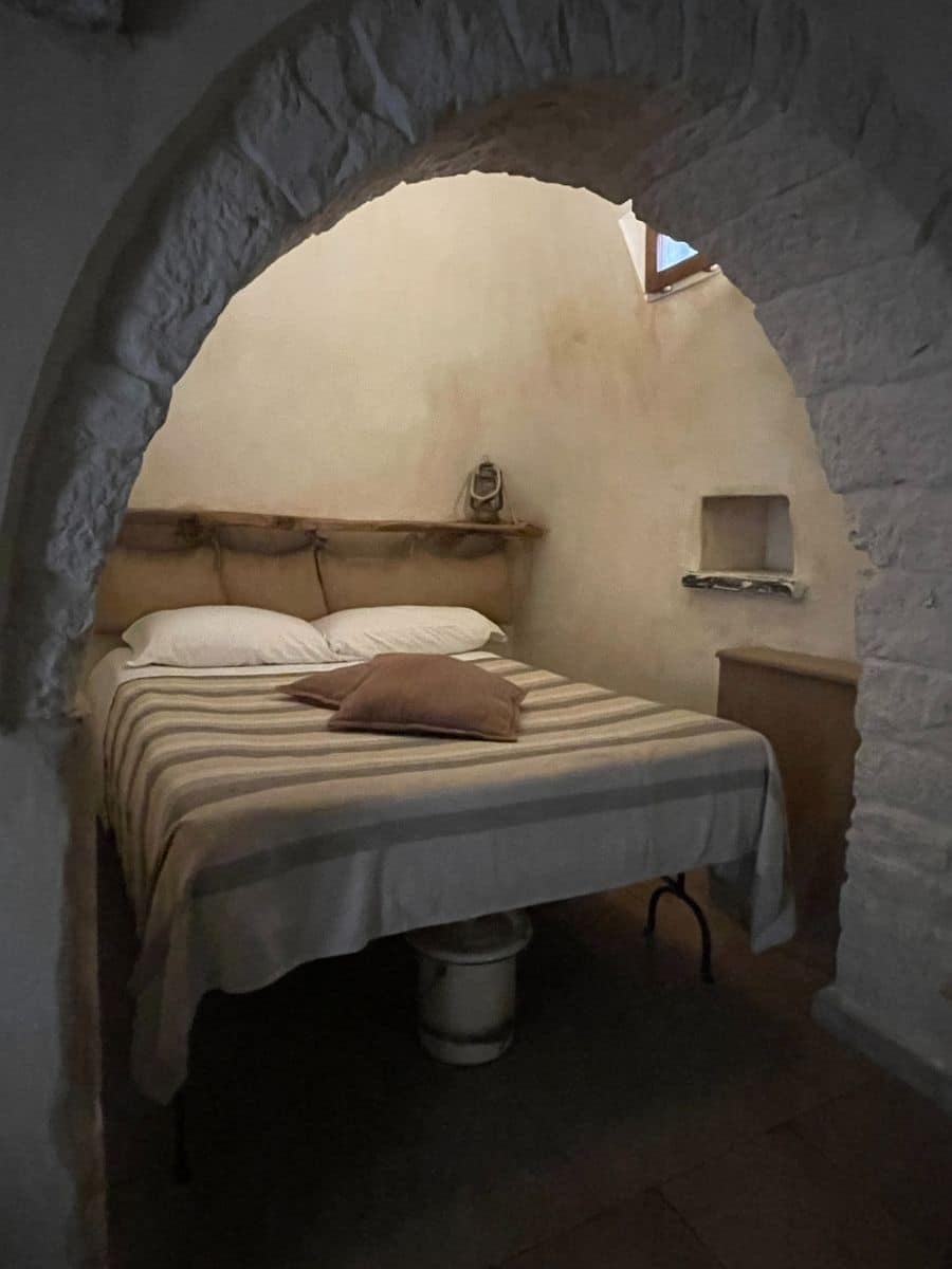A cozy bedroom inside a traditional Trullo house in Alberobello, Italy. The room is accessed through a rustic stone archway, leading to a bed with a striped beige and white bedspread and two pillows. A wooden shelf above the bed holds a vintage lantern, adding to the historical charm. A small built-in wall niche provides additional storage. A wooden cabinet stands to the right of the bed, and a small window near the ceiling allows soft natural light to enter. The space has an old-world, intimate feel, with its whitewashed stone walls and simple yet functional furnishings.