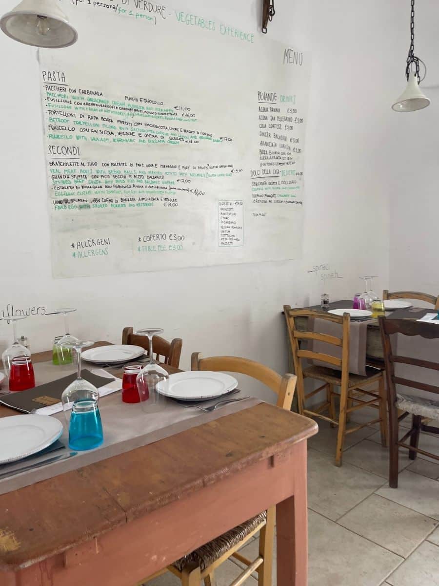 A charming rustic restaurant in Alberobello, Italy, with a simple and cozy atmosphere. The white walls feature a handwritten menu listing traditional Italian dishes such as pasta, secondi (main courses), and beverages. The wooden tables are set with white plates, silverware, and colorful inverted glasses in red, blue, green, and yellow, adding a playful touch to the setting. The mismatched wooden chairs contribute to the homey feel of the space. Hanging industrial-style lamps provide lighting. The overall ambiance is casual yet inviting, perfect for enjoying authentic local cuisine.