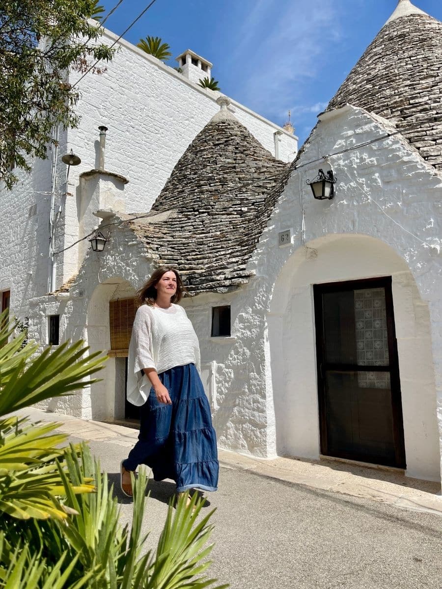 Melissa in a flowing white top and a tiered blue skirt walks gracefully past the traditional trulli houses of Alberobello, Italy. The trulli, with their iconic conical stone roofs and whitewashed walls, stand against a bright blue sky. Sunlight casts sharp shadows, highlighting the architectural details and rustic charm of the historic structures. A black lantern hangs near the entrance of one house, while a wooden shutter and small windows add character to the façade. Lush green plants frame the foreground, adding depth to the image.