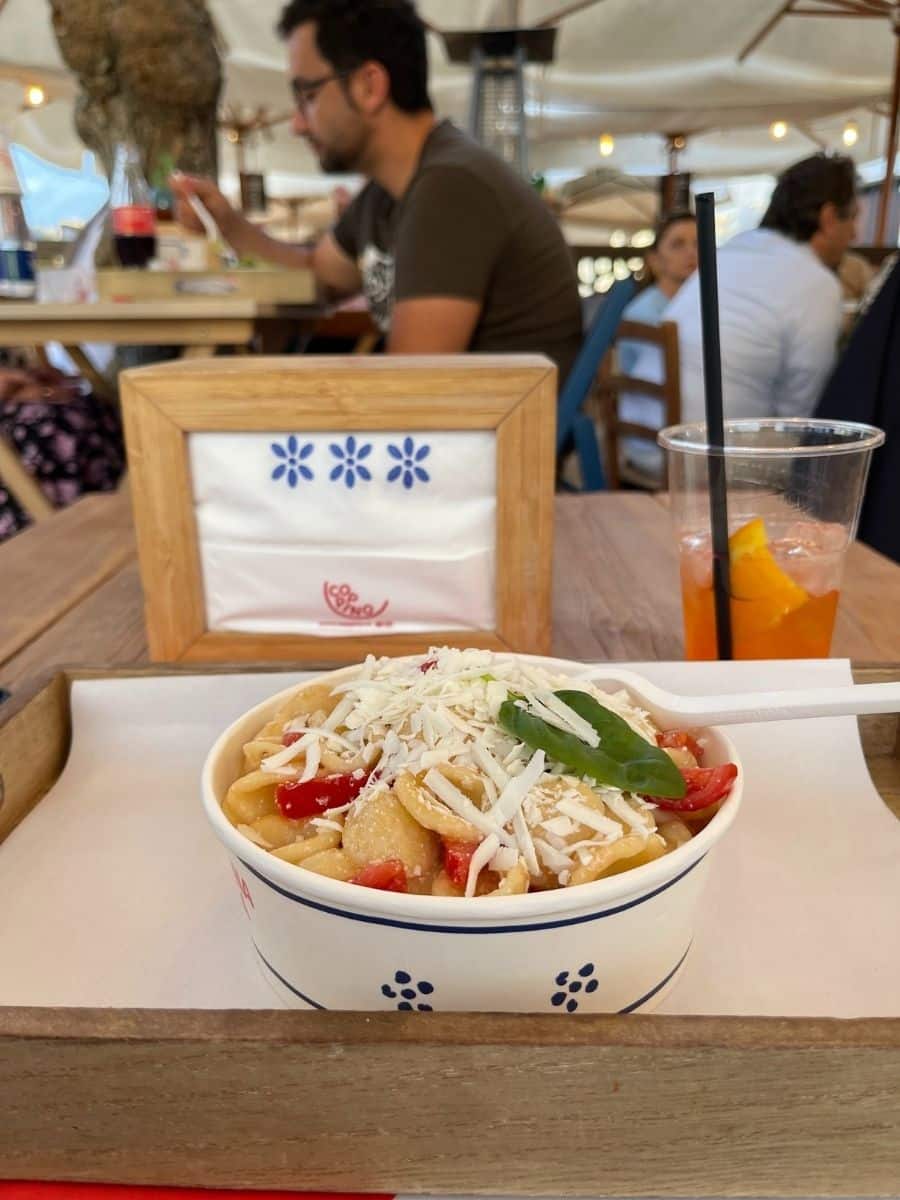 A bowl of freshly prepared orecchiette pasta, topped with grated cheese, cherry tomatoes, and a fresh basil leaf, sits on a wooden tray at an outdoor dining area. A white plastic spoon rests on the edge of the bowl. In the background, a napkin holder with a blue floral pattern and the restaurant's logo is visible. An orange Aperol Spritz with ice and a black straw sits nearby. Other diners are engaged in conversation under a canopy, creating a lively atmosphere at the eatery.