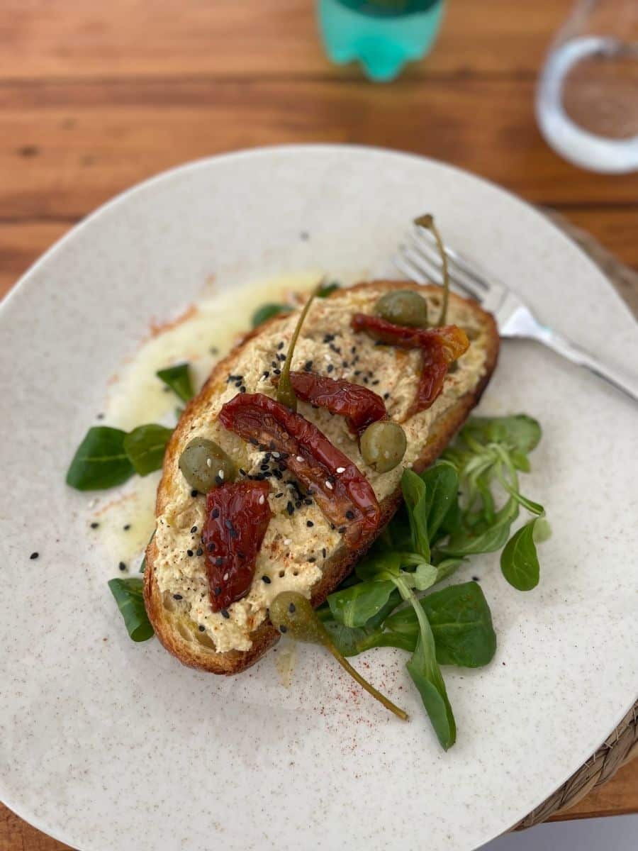 A beautifully plated dish featuring a slice of rustic toasted bread topped with a creamy spread, sun-dried tomatoes, green olives, caper berries, and black sesame seeds. The toast is placed on a bed of fresh green salad leaves and is lightly dusted with paprika. A silver fork rests beside the toast on a speckled ceramic plate, set on a wooden table with a partially visible water glass and a green bottle in the background.
