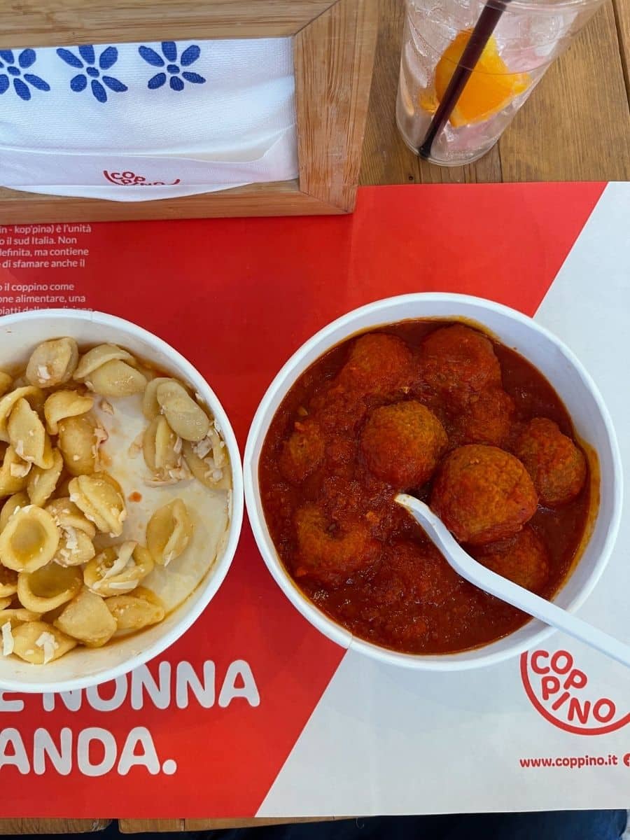 A meal featuring two bowls placed on a red and white placemat with the text 'Coppino' and 'Nonna'. One bowl contains orecchiette pasta with grated cheese, while the other is filled with meatballs in a rich tomato sauce. A white plastic spoon rests in the meatball bowl. In the background, a glass with an orange slice and a black straw, along with a wooden napkin holder decorated with blue floral designs, are visible on a wooden table.