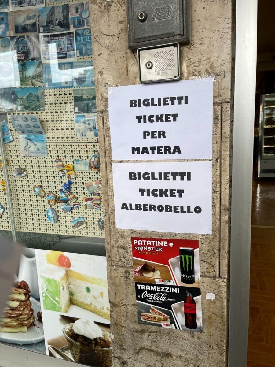 A wall outside a ticket-selling shop with two printed signs taped to it, offering tickets for travel to Matera and Alberobello. The signs read 'BIGLIETTI TICKET PER MATERA' and 'BIGLIETTI TICKET ALBEROBELLO'. Below them, two colorful advertisements promote snacks and drinks, including Monster Energy and Coca-Cola Zero Sugar, alongside images of tramezzini sandwiches and chips. The window display in the background features postcards and images of tourist destinations, as well as cakes and desserts.