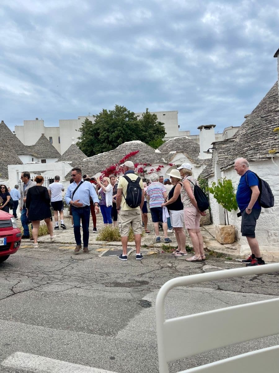 A group of tourists exploring the charming streets of Alberobello, Italy, surrounded by traditional trulli houses with their iconic conical stone roofs. The visitors, dressed in casual summer attire with backpacks and hats, appear engaged in sightseeing. Some are taking photos, while others are conversing. The narrow street is lined with whitewashed trulli, some decorated with vibrant red bougainvillea. The pavement shows visible cracks, adding to the rustic character of the scene. The sky is overcast, casting a diffused light over the picturesque setting.