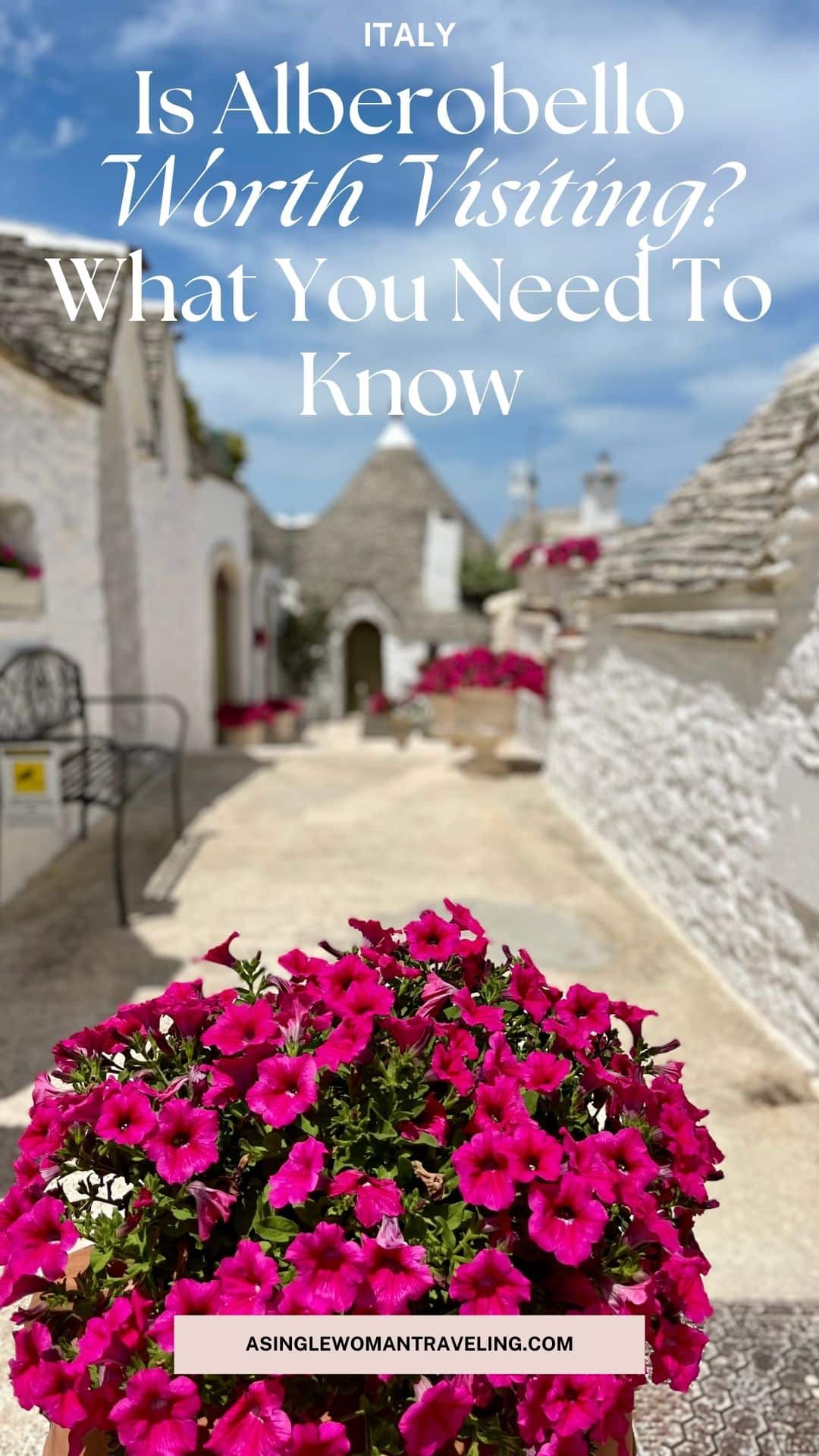 A picturesque street in Alberobello, Italy, lined with traditional white trulli houses with conical roofs. A vibrant cluster of pink flowers in the foreground adds charm to the sunlit stone pathway. The text overlay reads, "Is Alberobello Worth Visiting? What You Need to Know," with the website ASINGLEWOMANTRAVELING.COM displayed at the bottom.
