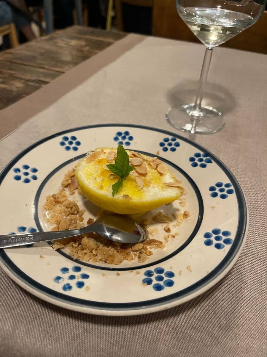 A beautifully plated lemon dessert served on a rustic ceramic plate with blue floral patterns. The dessert consists of a halved lemon filled with a sorbet-like treat, topped with toasted almond slices and a fresh mint leaf. Crumbled biscuits surround the lemon, adding texture. A spoon rests on the plate, ready to dig in. In the background, a glass of white wine sits on a linen-covered wooden table, creating a cozy and inviting dining atmosphere.