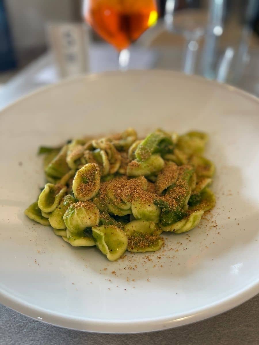 A plate of freshly made orecchiette pasta coated in a vibrant green pesto sauce, garnished with toasted breadcrumbs. In the blurred background, a glass of Aperol Spritz and other dining elements add to the inviting dining atmosphere.