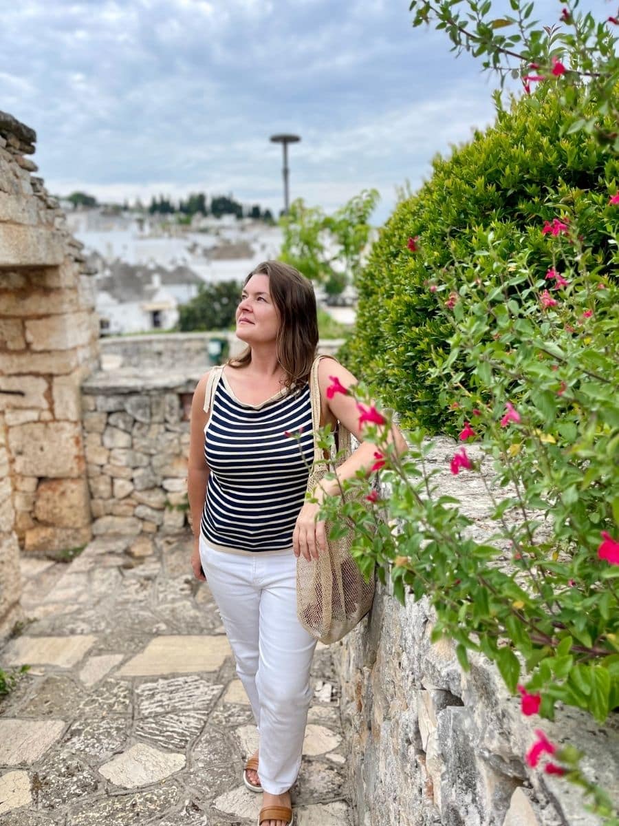 A woman in a striped sleeveless top and white pants walks along a stone pathway in Alberobello, Italy. She carries a net bag and looks up with a relaxed and content expression. The background features the iconic white trulli houses with conical roofs, while lush greenery and pink flowers frame the foreground. The sky is overcast, adding a soft, diffused light to the scene.