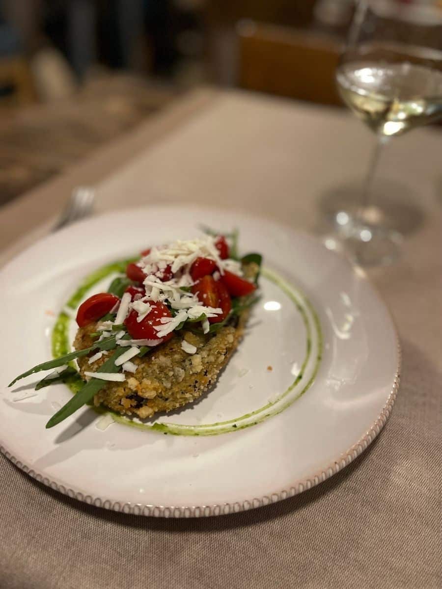 A beautifully plated dish featuring a crispy, breaded vegetable patty topped with fresh cherry tomatoes, arugula, and grated cheese. The dish is elegantly presented on a white plate with a decorative green sauce drizzle. In the blurred background, a glass of white wine is visible, adding to the sophisticated dining ambiance. The table setting includes a beige tablecloth, creating a warm and inviting atmosphere.