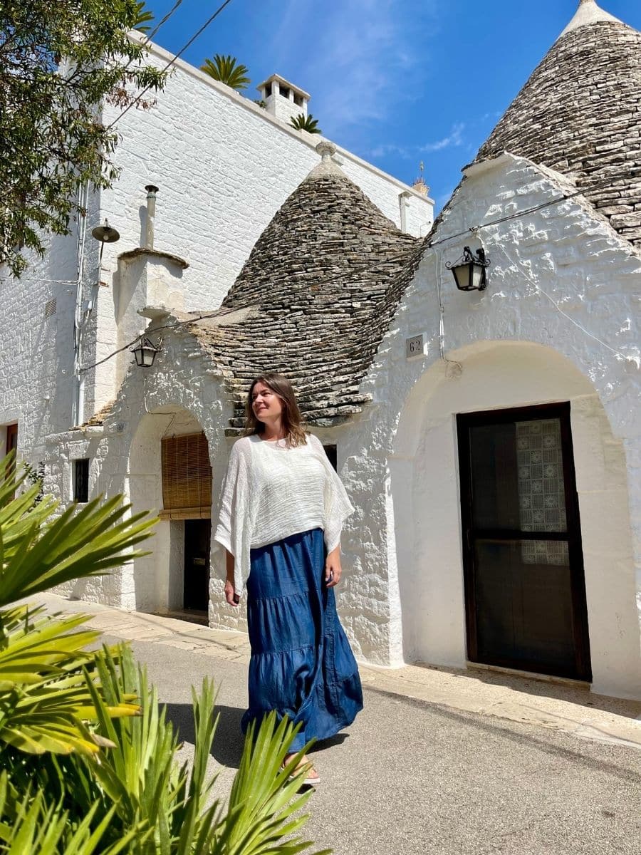 A woman with light skin and brown hair walks barefoot on a sunlit street in Alberobello, Italy, surrounded by traditional trulli houses. She wears a flowing white blouse with wide sleeves and a long blue skirt. She gazes into the distance with a serene expression. The bright blue sky and white stone buildings create a picturesque Mediterranean backdrop, with lush green palm leaves in the foreground.