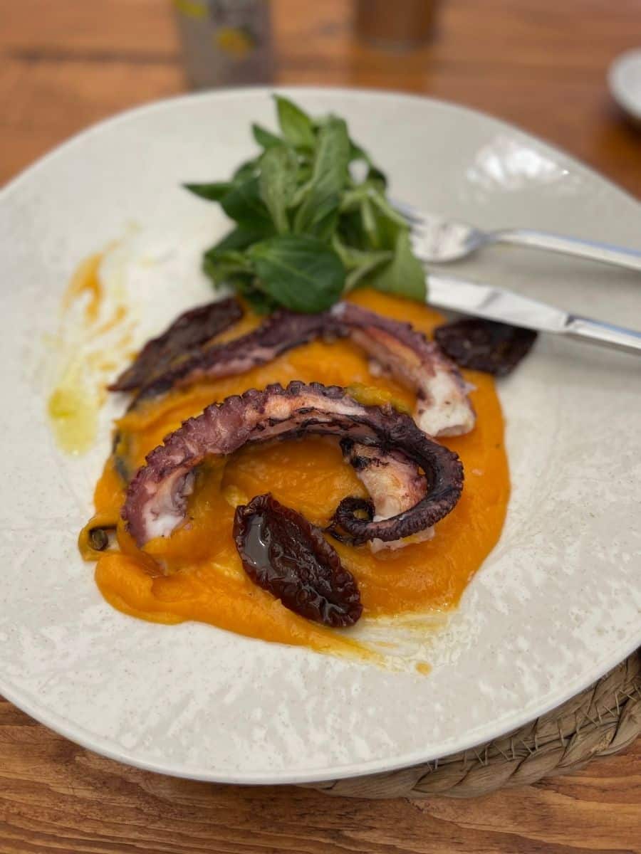 A gourmet dish featuring grilled octopus with a slightly charred exterior, served on a bed of vibrant orange purée. The dish is garnished with sun-dried tomatoes, fresh green salad leaves, and drizzled with olive oil. A fork and knife rest on the side of the white ceramic plate, which is placed on a rustic wooden table. The background is slightly blurred, suggesting an outdoor dining setting.