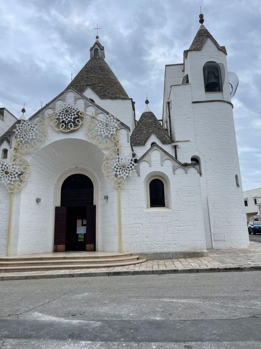 A historic white church in Alberobello, Italy, featuring the town’s signature trulli-style architecture with conical stone roofs. The façade is adorned with intricate decorative lights, suggesting a festival or special occasion. A bell tower rises on the right side, while the open wooden doors invite visitors inside. The sky is overcast, casting a soft, diffused light over the scene.