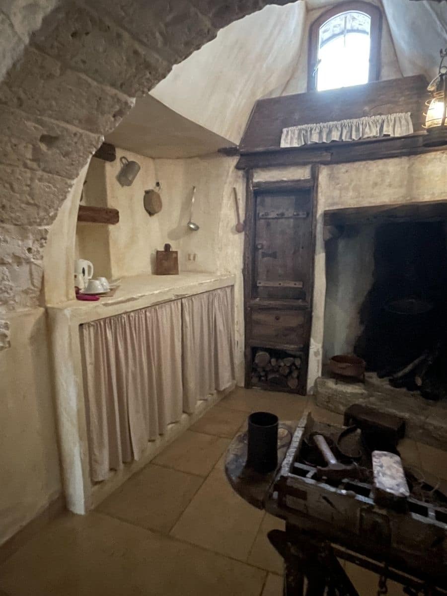 A rustic interior of a traditional trullo house in Alberobello, Italy. The space features stone walls and an arched ceiling, with a small window allowing natural light in. The kitchen area has a simple countertop with a fabric curtain covering the storage space below, and various old-fashioned kitchen tools hanging on the wall. To the right, there is a fireplace with a wooden cupboard above it for additional storage. A wooden worktable with antique tools sits in the foreground, adding to the historical charm of the room.