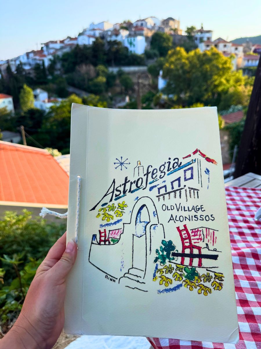 Hand holding a menu from Astrofegia Restaurant in Old Village Alonissos, featuring a hand-drawn illustration with whitewashed steps, red chairs, and Greek-style architecture, set against a backdrop of hillside houses and greenery.