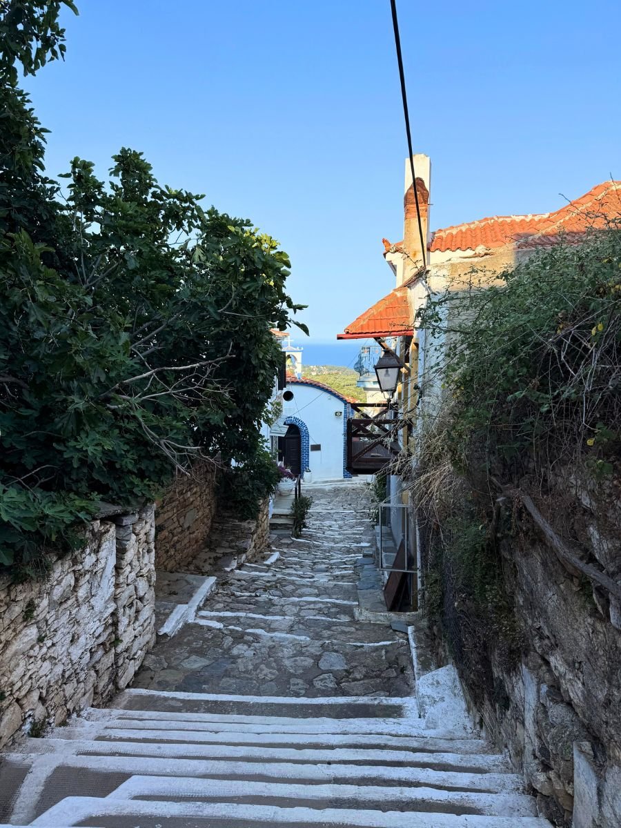 Stone stairway flanked by stone walls and lush greenery, leading to a small white church with a blue arched doorway, set against a backdrop of red rooftops and a view of the sea in the distance.