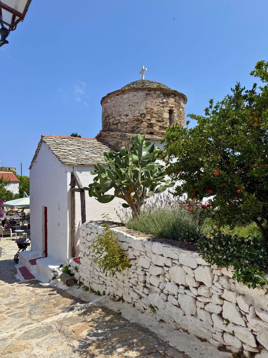 Old stone chapel with a rounded dome and white cross on top, surrounded by whitewashed walls, cacti, and flowering plants in Alonissos, Greece.