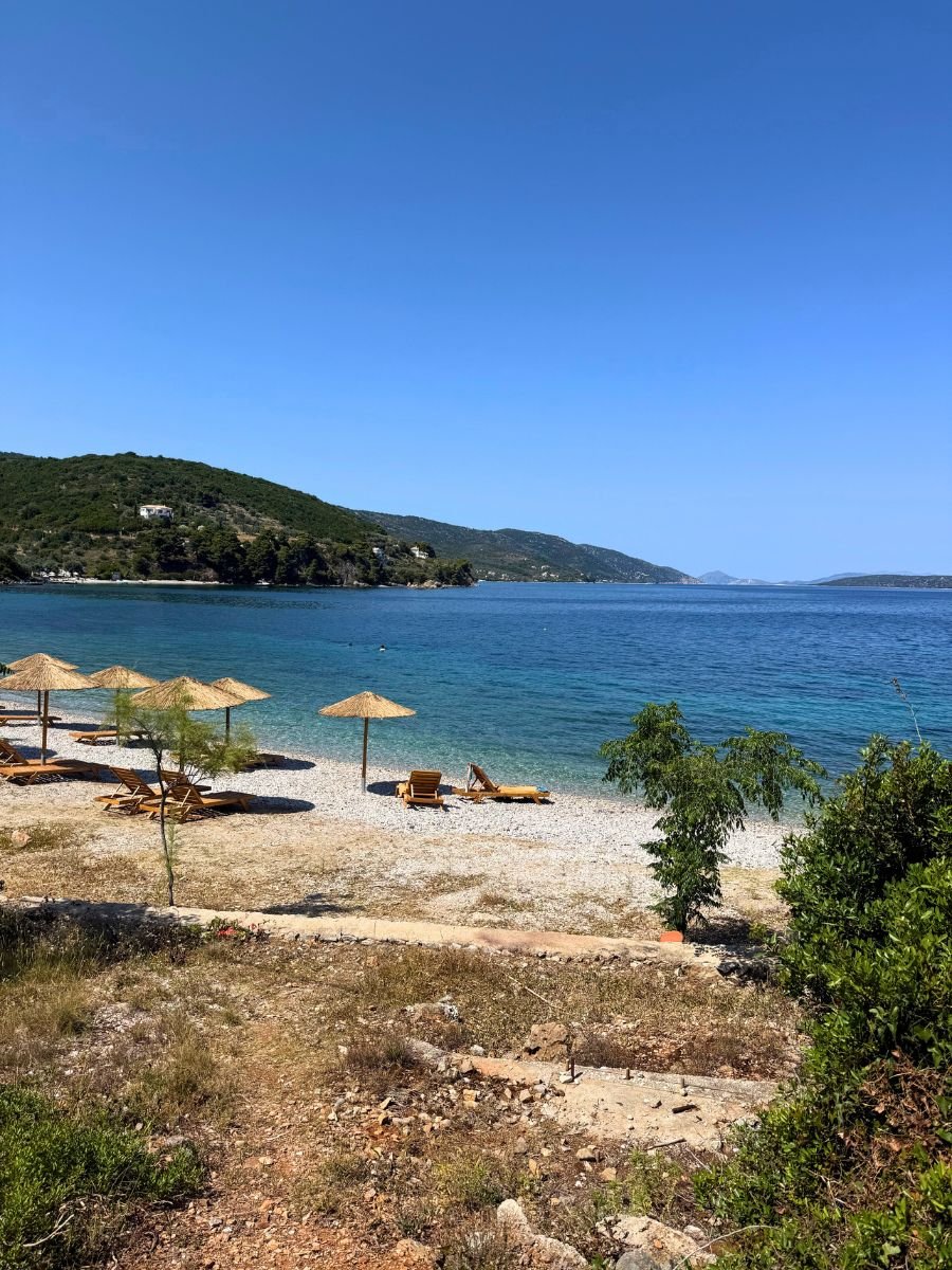Secluded beach in Alonissos with straw umbrellas, wooden sunbeds, and turquoise water backed by lush green hills under a clear blue sky.