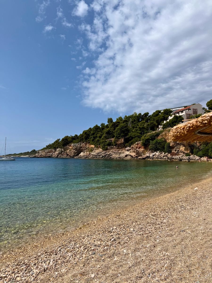 Tranquil pebbled beach in Alonissos, Greece, with crystal-clear blue water, a lone swimmer, rocky coastline covered in pine trees, and a sailboat anchored in the distance under a partly cloudy sky.