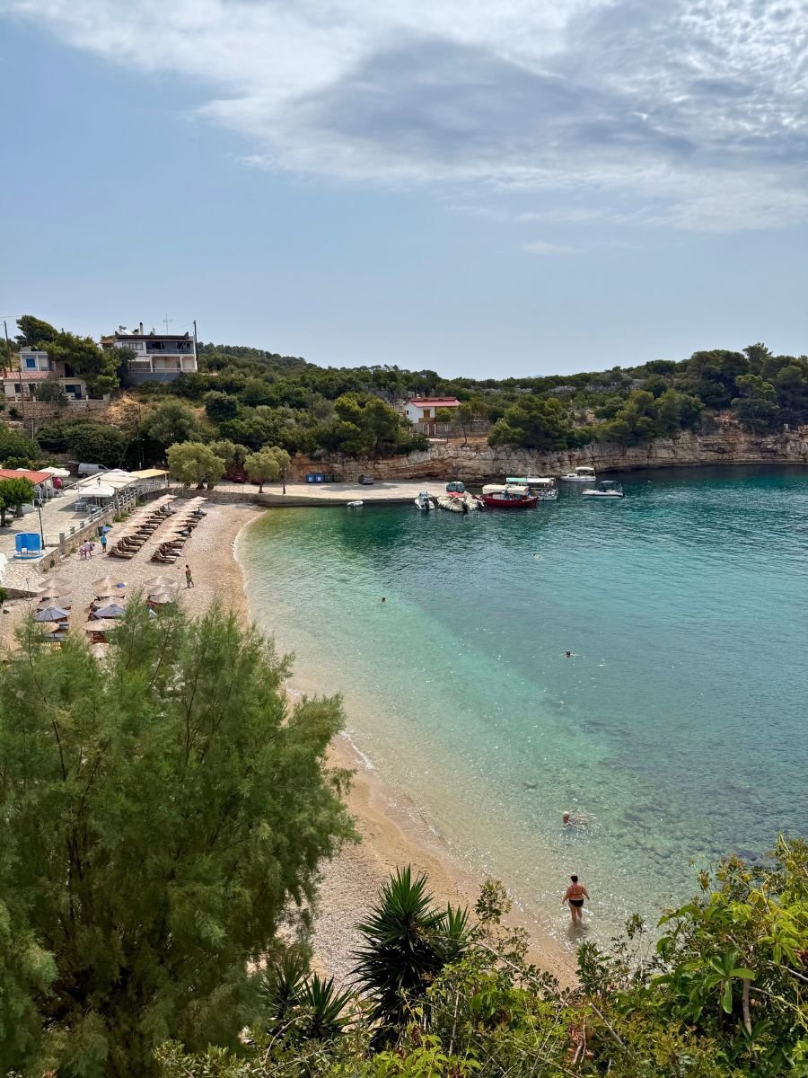 Scenic beach cove in Alonissos, Greece, with crystal-clear turquoise waters, a few swimmers, and sunbeds lined along a small pebbled shore, surrounded by lush greenery and boats docked at a quiet pier.