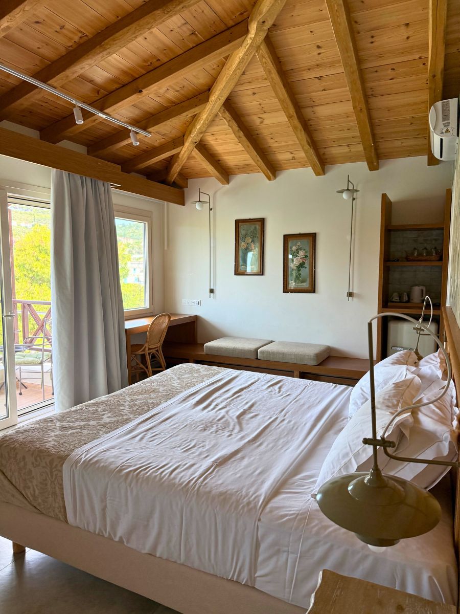 Cozy bedroom with wooden ceiling, neatly made bed, and a balcony door letting in natural light, in a rustic-style accommodation in Alonissos, Greece.