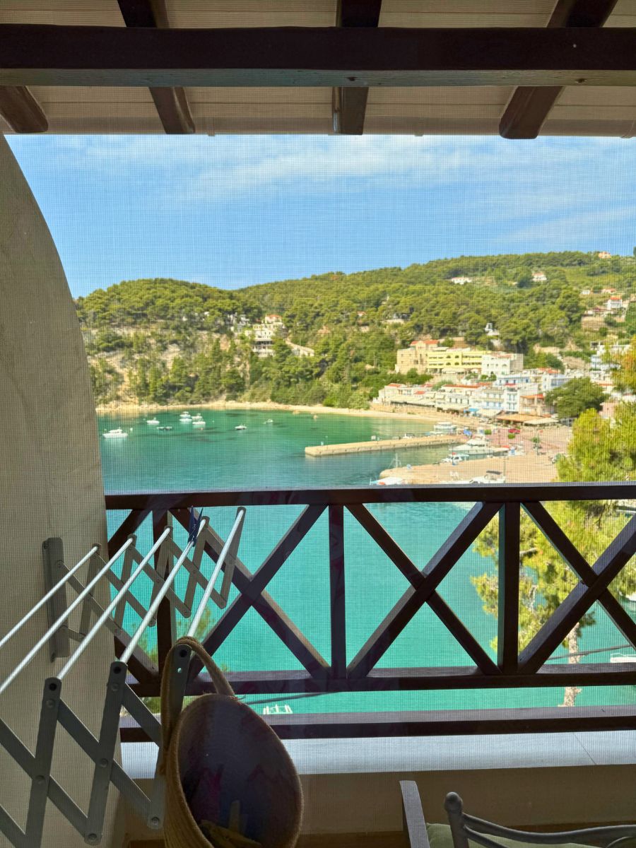 Scenic view of a turquoise bay and coastal village in Alonissos, Greece, as seen through a balcony with wooden railing and drying rack.