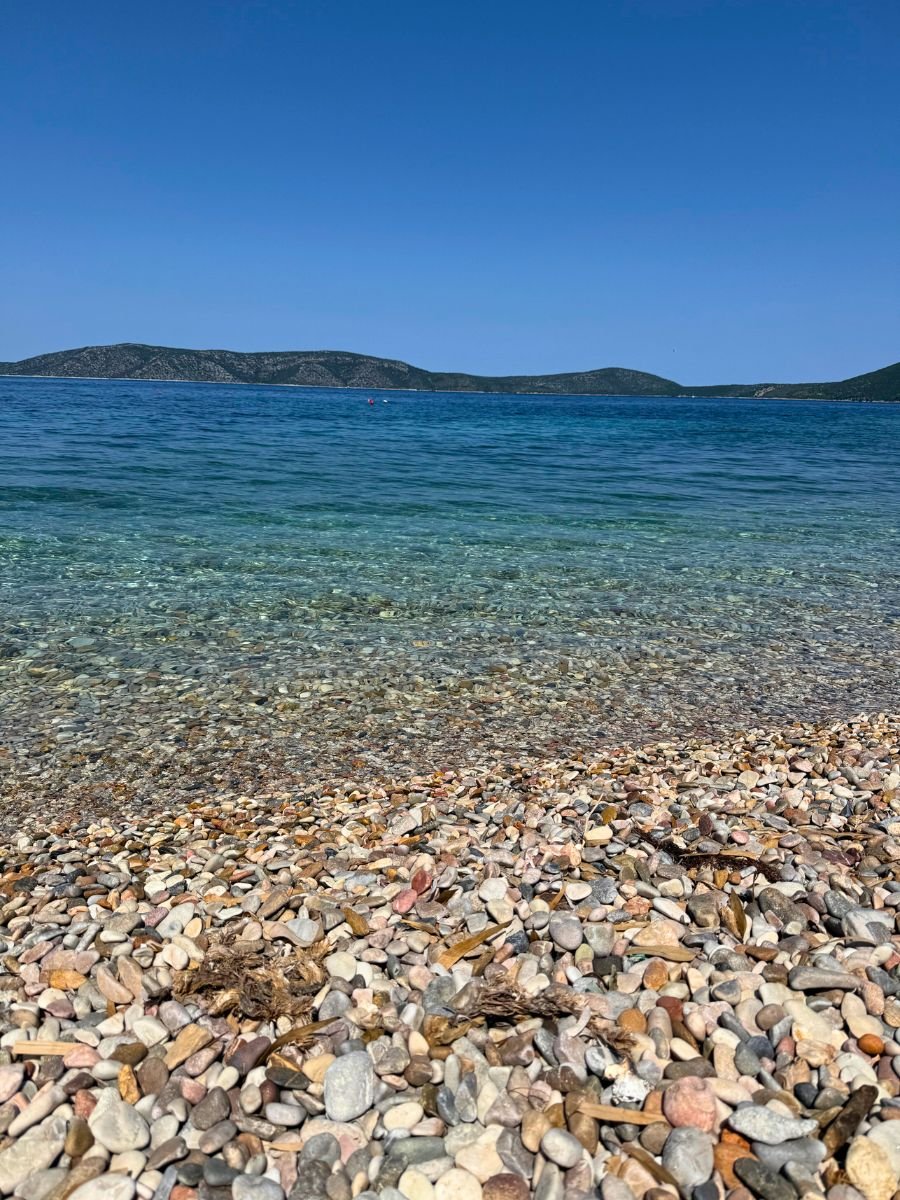 Pebble beach on Alonissos island with clear turquoise water gently lapping the shore, and distant green hills under a cloudless blue sky.