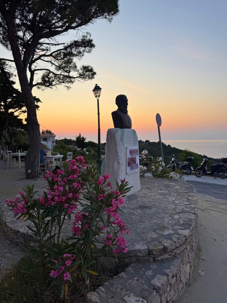 Statue of a man atop a stone pedestal surrounded by blooming pink flowers at sunset in Alonissos, with a row of scooters parked nearby and a glowing horizon over the Aegean Sea.