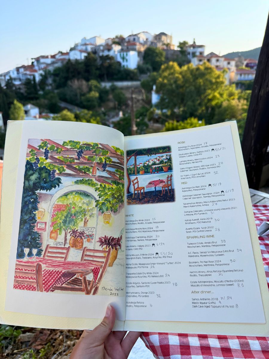 Hand holding a beautifully illustrated wine menu with colorful paintings of a taverna, with the hillside village of Alonissos in the background under soft evening light.