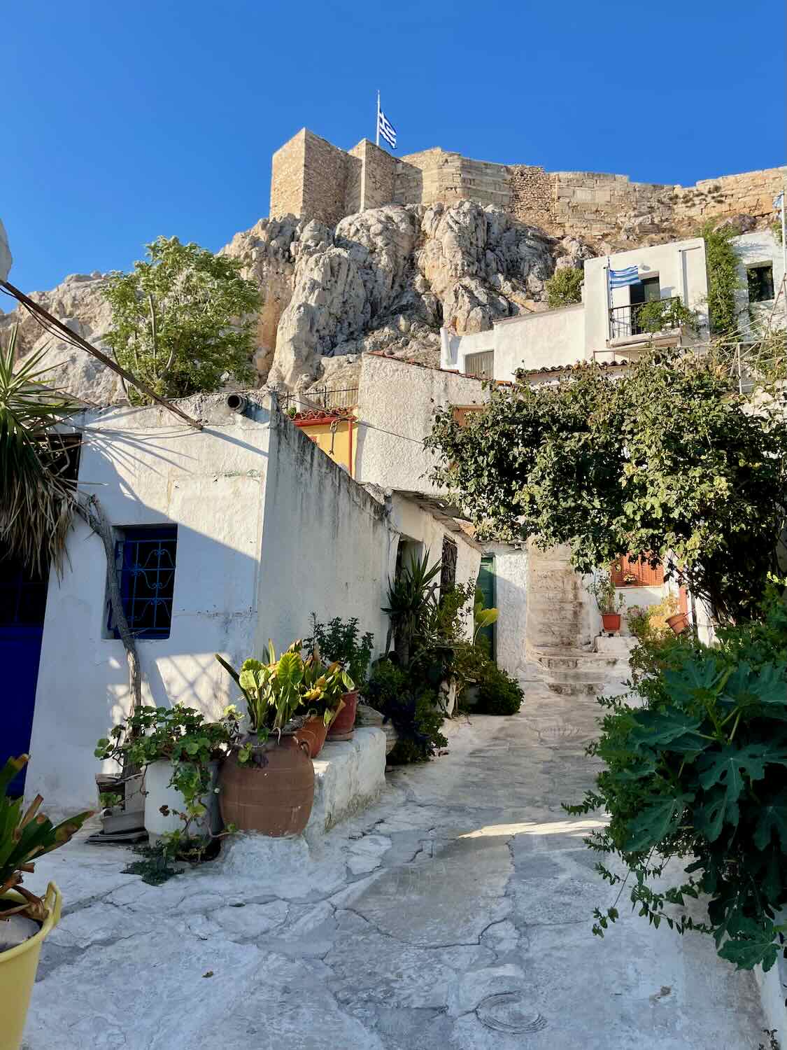 A quaint cobblestone pathway leads up through a traditional Greek village in Athens in the off season, with white-washed houses adorned with potted plants and a clear blue sky above. In the background, the imposing structure of an ancient fortress sits atop a rugged hill, overseeing the village.
