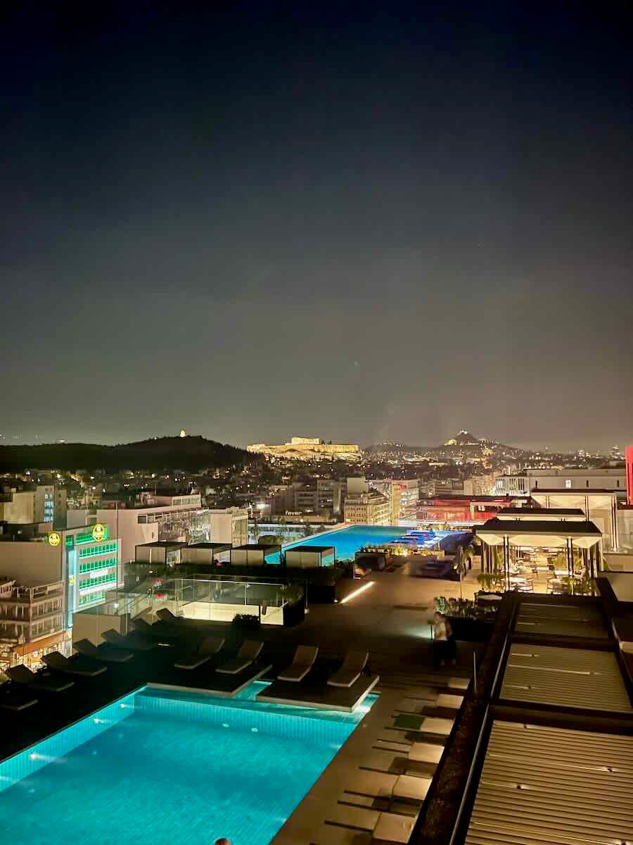 A stunning rooftop pool view in Athens at night, with the illuminated Acropolis glowing in the background, blending modern luxury with ancient history.