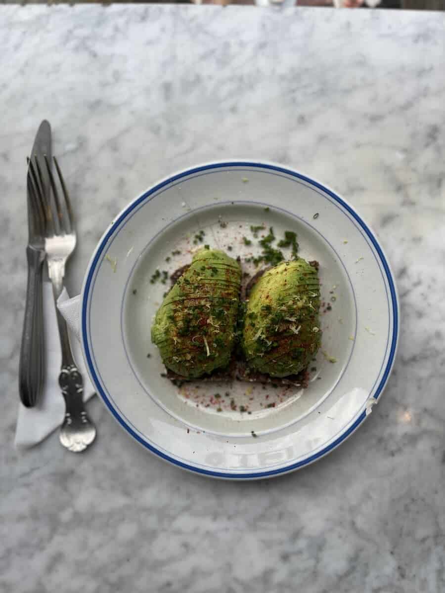 Healthy breakfast option at Atelier September featuring two avocado toasts heavily garnished with herbs and spices, presented on a vintage plate with a blue rim, set on a marble table.