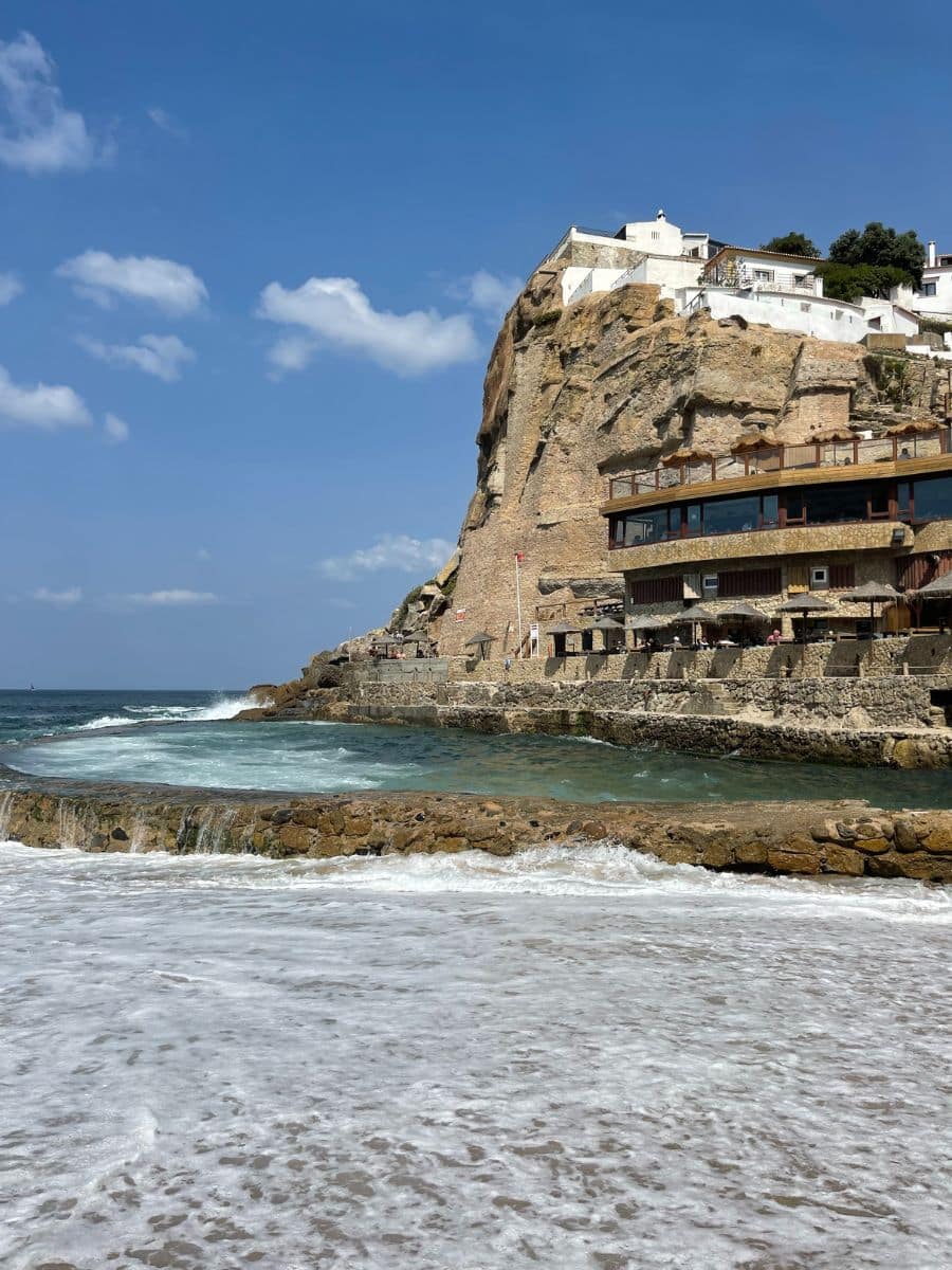 Picturesque view from the beach of Azenhas do Mar village perched on dramatic cliffs, waves gently breaking onto the sandy shore.