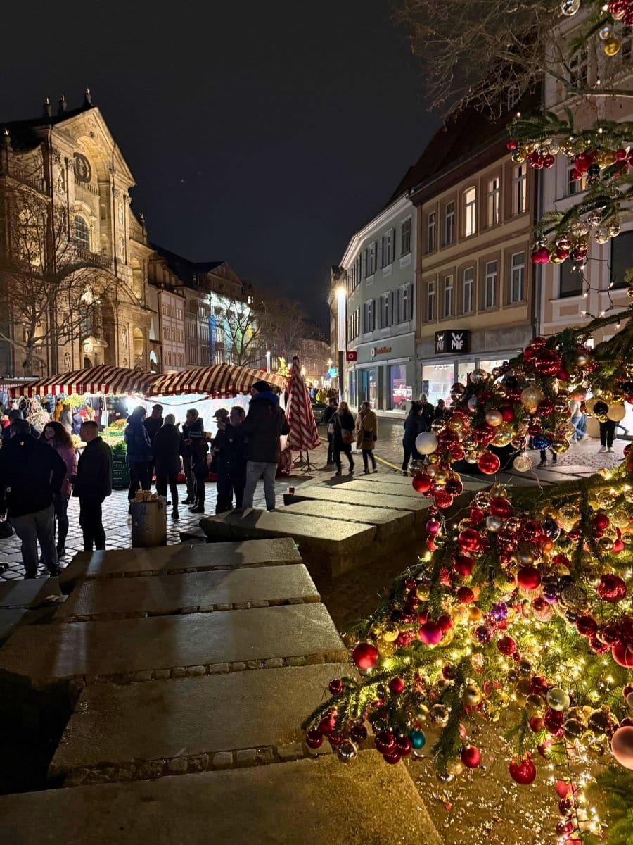 A lively Christmas market scene in Bamberg at night with crowds gathering near red-and-white striped booths and a decorated Christmas tree glowing in the foreground.