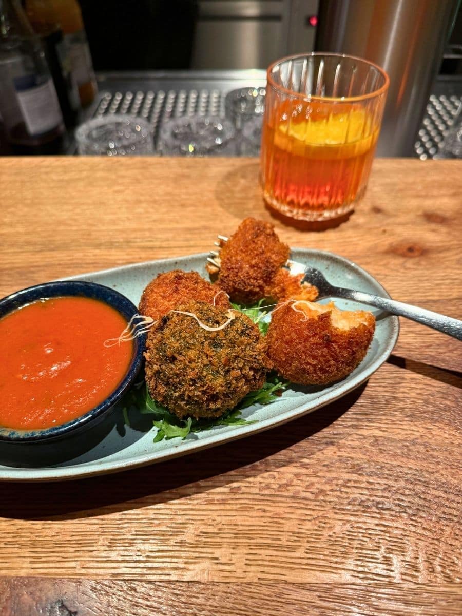 Close-up of a bar counter with a plate of crispy croquettes served with red dipping sauce, next to a glass of orange-colored cocktail.