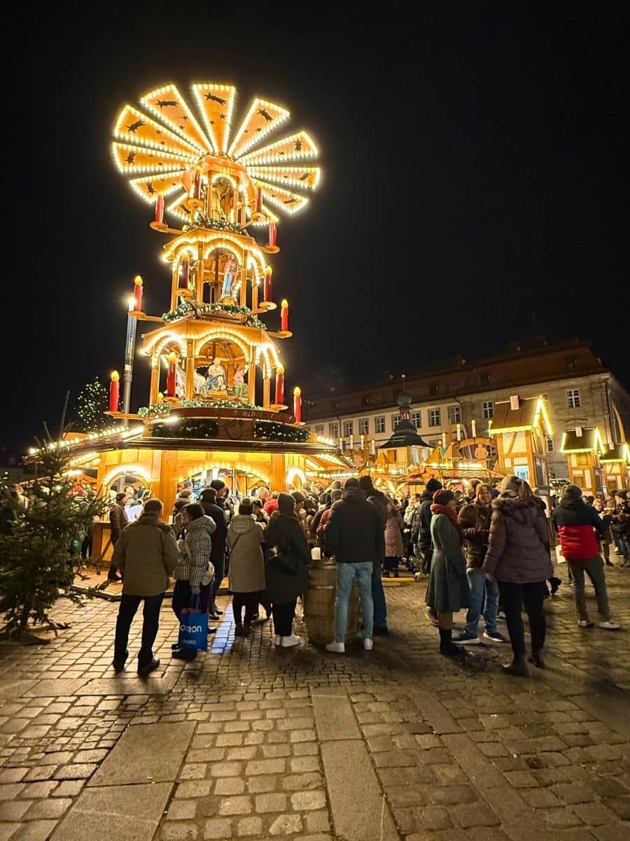 Crowds gathered around a towering illuminated Christmas pyramid structure at Bamberg’s festive night market, surrounded by glowing wooden booths and holiday cheer.
