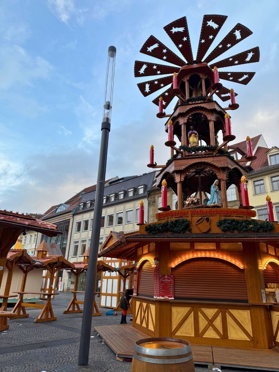 Tall wooden Christmas pyramid structure with candles and nativity figures in Bamberg’s holiday market, surrounded by closed market stalls.