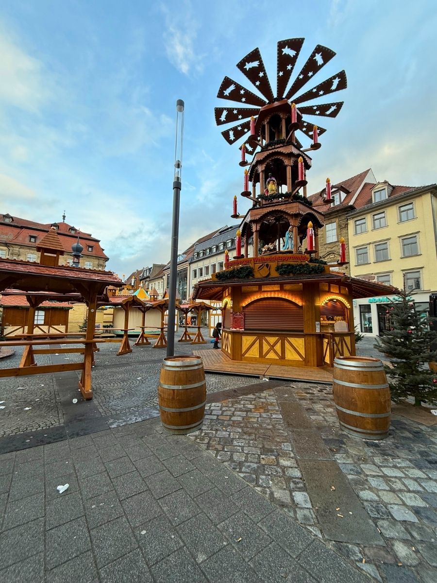 Large wooden Christmas pyramid with candles and nativity figures at a festive market in Bamberg’s town square, surrounded by half-timbered stalls and barrels.