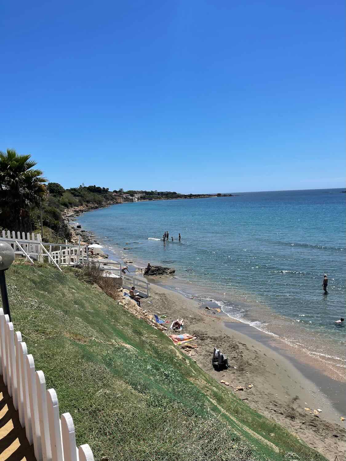 A beach scene with clear blue water and a sandy shore. There are a few people enjoying the water and sunbathing on the sand. The sky is bright blue with no clouds.