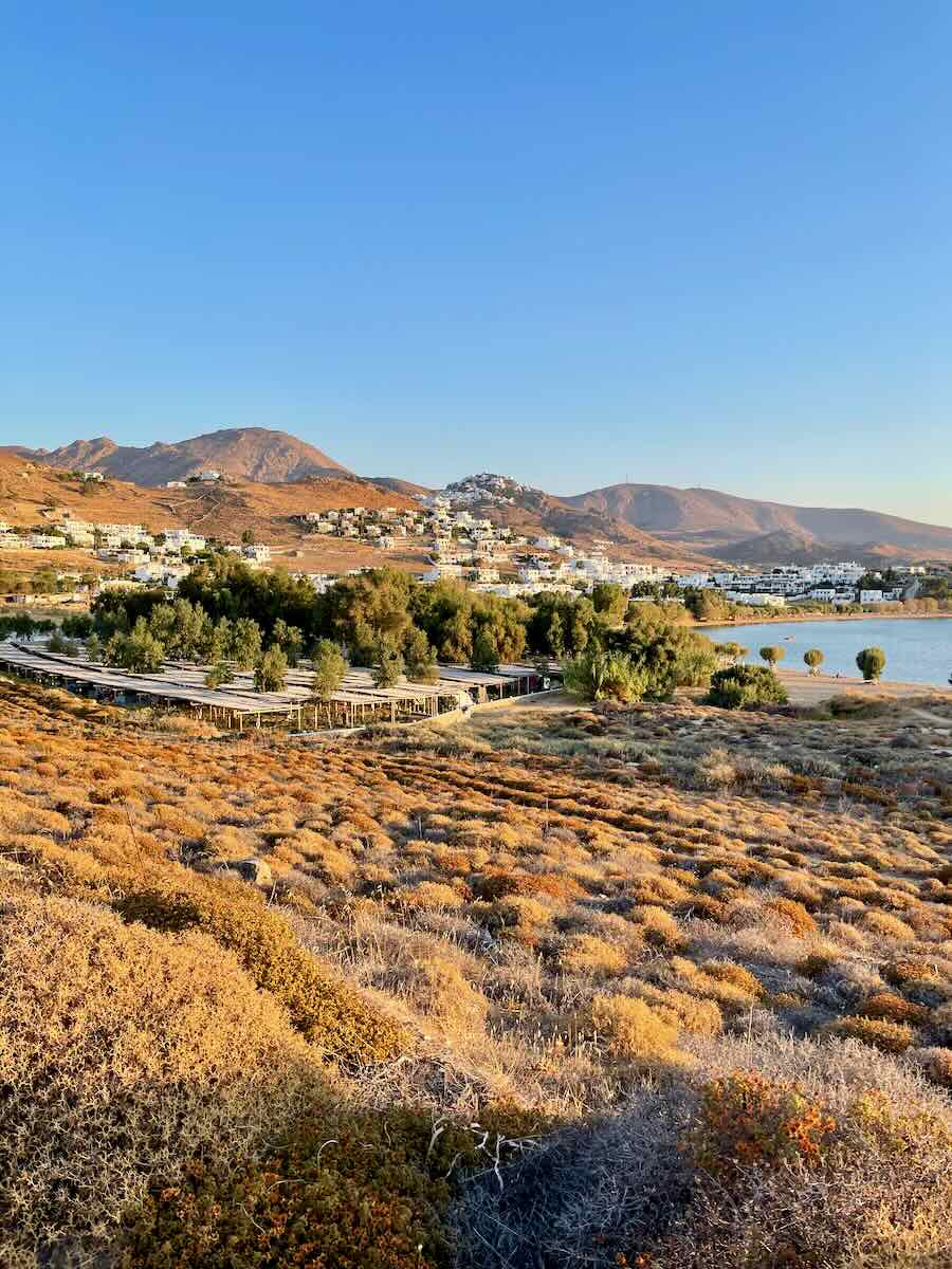 The image shows a sunlit, arid landscape with dry shrubs in the foreground, leading to a coastal area lined with trees and buildings. In the distance, white houses dot the hillside under a clear blue sky. The scene captures a rugged yet picturesque Mediterranean setting, likely near a beach or camping area, with mountains visible in the background.
